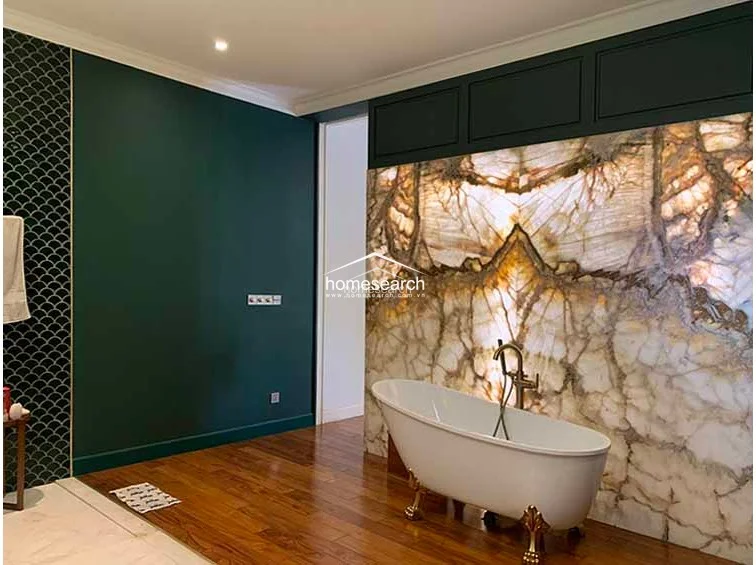 This bathroom area highlights a dramatic design contrast, pairing a deep green painted wall with an impressive, illuminated onyx stone wall that serves as a focal point for the clawfoot tub. The combination of warm wood flooring and elegant brass hardware reflects a high level of maintenance and a refined, artistic architectural style.