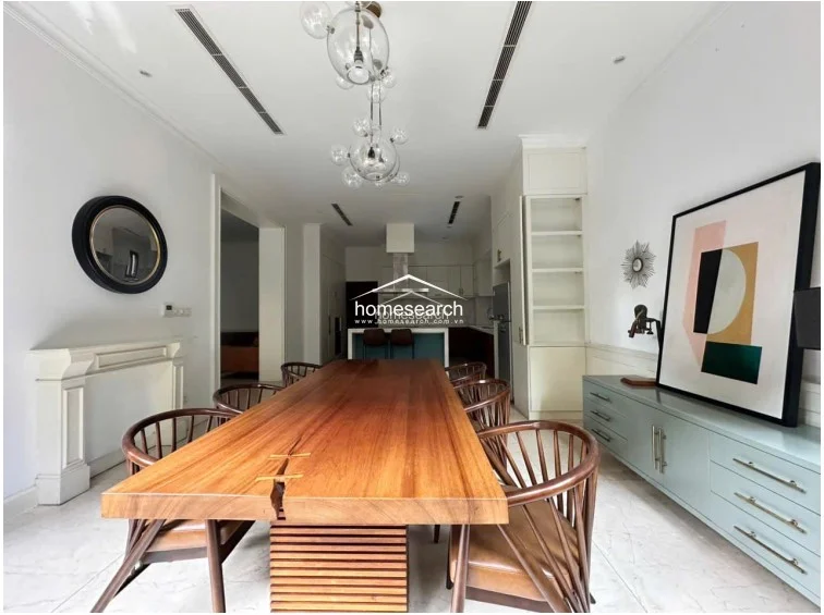 This dining area is defined by a massive, solid wood slab table and a unique, multi-globe pendant light fixture. The open-concept space seamlessly connects to a kitchen in the background and features built-in white cabinetry, marble floors, and a decorative fireplace mantel.