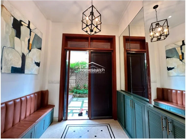 This entryway serves as a sophisticated mudroom or foyer, featuring built-in green cabinetry with integrated leather bench seating. The space is highlighted by a decorative geometric pendant light, large wall mirrors that expand the visual depth, and a direct view out to a private, paved garden area.