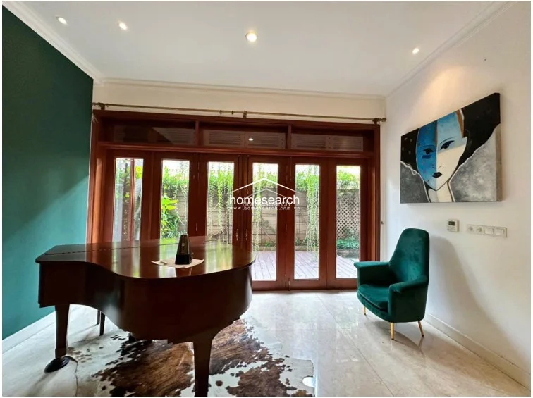 This music room is anchored by a grand piano set upon a stylish cowhide rug, with polished marble flooring reflecting the natural light from the expansive floor-to-ceiling wooden glass doors. The space features a bold dark green accent wall that contrasts beautifully with the neutral tones and provides a direct, seamless flow to the outdoor wooden deck.