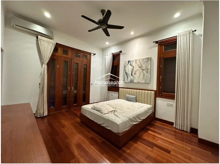 This bedroom features warm hardwood flooring and a central bed with a neutral upholstered headboard. The room is well-lit by natural light from multiple windows and features a ceiling fan, wall-mounted AC unit, and elegant wooden framed doors that likely lead to an outdoor area.