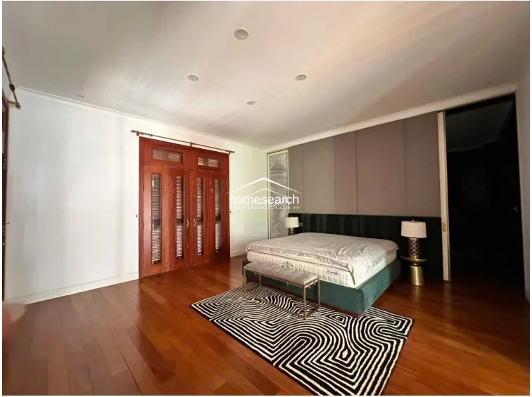 This bedroom is defined by a sophisticated color palette, featuring a dark upholstered headboard against a grey paneled accent wall and rich hardwood floors. A stylish geometric area rug anchors the bed, while the room is illuminated by recessed lighting and elegant bedside lamps.