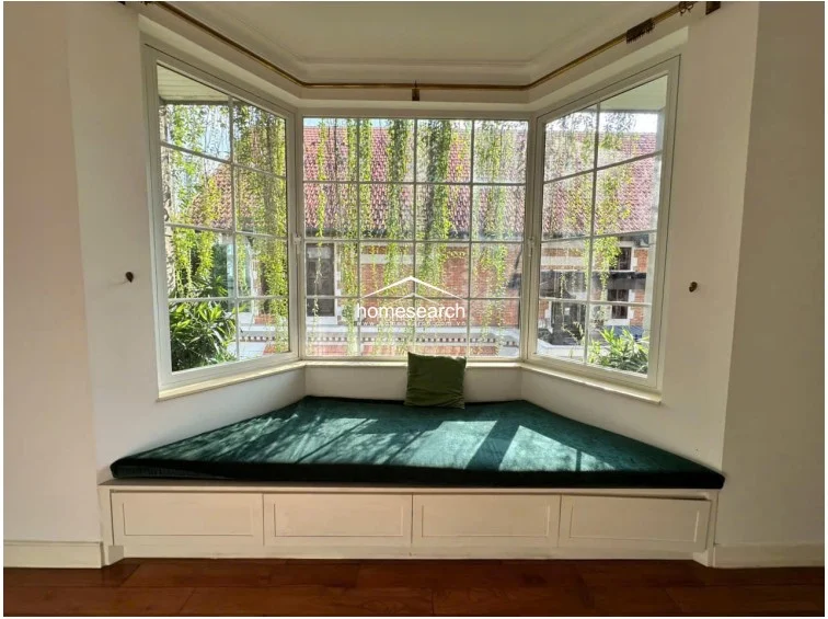This charming bay window area serves as a cozy reading nook, featuring a custom-built bench with integrated storage drawers underneath. The large, multi-paned windows offer a picturesque view of greenery and traditional roof tiles, flooding the space with soft natural light.