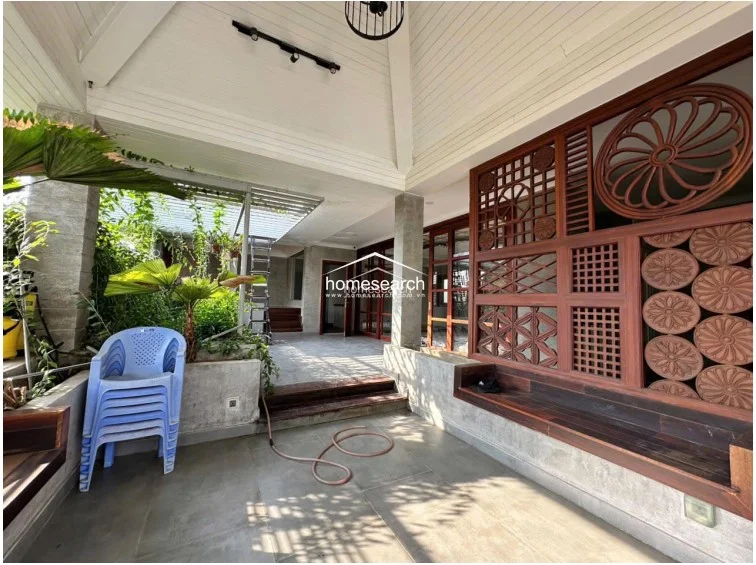 This expansive covered patio serves as a seamless indoor-outdoor transition space, featuring large tiled flooring and a high vaulted ceiling. It is adorned with intricate wooden lattice panels and a built-in bench, offering a lush view of the adjacent garden and greenery.