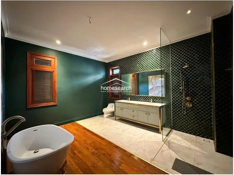 This luxurious bathroom features a freestanding soaking tub set against a rich green wall, complemented by warm wood flooring. The walk-in shower area is encased in glass and adorned with dark green, scale-patterned tiles, creating a sophisticated and high-end aesthetic.