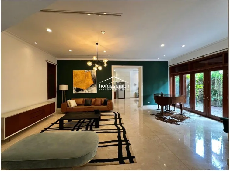 The spacious living area is defined by a striking dark green accent wall and large wooden-framed glass doors that provide ample natural light and views of the greenery outside. It features a modern chandelier, a sleek leather sofa, and a large patterned area rug, all set against polished light tile flooring.
