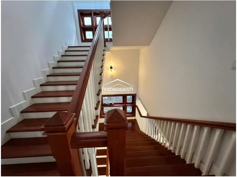 This staircase area features a classic design with rich, dark wood treads and a contrasting white balustrade. The space is well-lit by a modern wall sconce and natural light filtering through small, grid-patterned wooden windows, creating an elegant and well-maintained transition between floors.