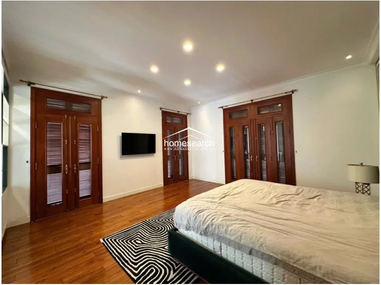 This spacious bedroom is characterized by warm wooden flooring and multiple sets of French-style wooden doors that provide access to the exterior. The room is minimalist in decor, featuring a large bed and a wall-mounted television, with recessed ceiling lighting providing a soft, ambient glow.
