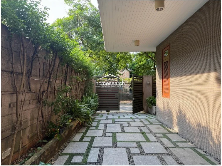 The exterior entrance features a private driveway paved with large stone slabs and decorative gravel, bordered by a lush vertical garden and mature trees. The property is secured by a modern metal gate, and the exterior wall is finished with textured horizontal cladding, presenting a well-maintained and contemporary facade.