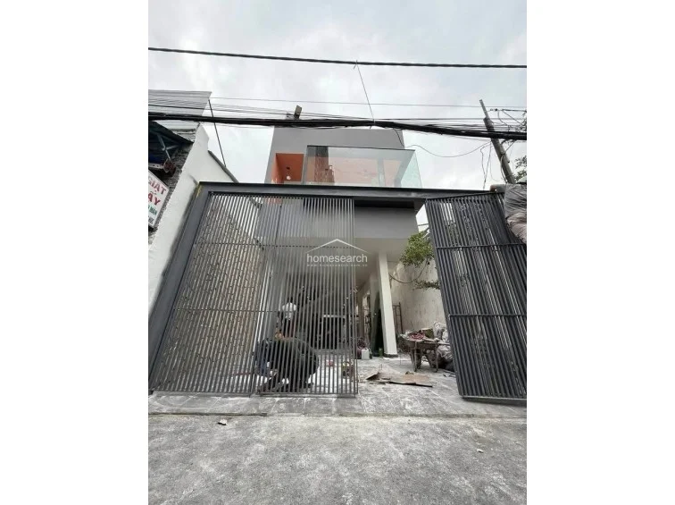 This exterior view shows a modern multi-story property currently undergoing construction or renovation, featuring a tall, vertical-slat metal gate for security. The facade incorporates a minimalist grey finish with a prominent glass-enclosed balcony on the upper level, suggesting a contemporary architectural style.