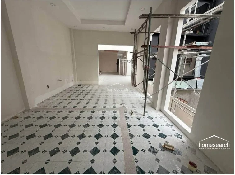 This room showcases a distinct decorative flooring pattern with diamond-shaped dark tiles set against a lighter background, adding a classic touch to the otherwise modern, unfinished space. The area is illuminated by natural light from a large side window and features a recessed tray ceiling, highlighting the ongoing interior finishing work.