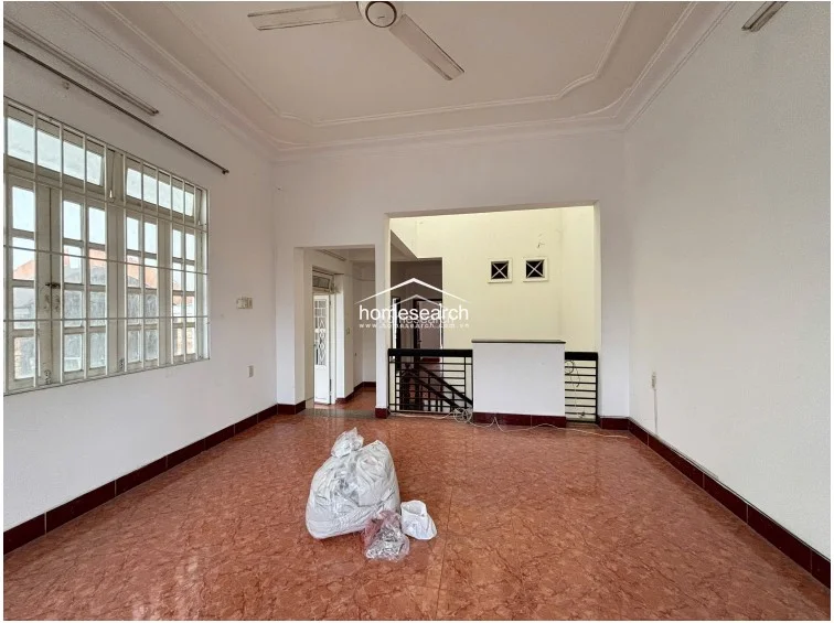 This bedroom or multi-purpose room is finished with reddish-brown patterned tile flooring and white walls with a dark baseboard. It features a ceiling fan, a large window with security bars, and an open doorway leading to a landing area with a staircase.