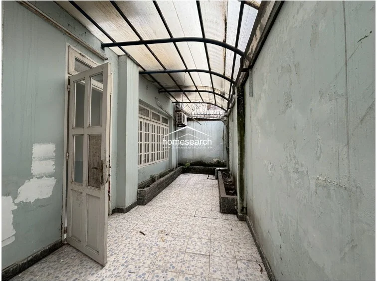This side passage or secondary outdoor area is covered by a translucent corrugated roof, providing a sheltered walkway or utility space. The walls show significant signs of wear and peeling paint, and the area includes built-in concrete planters along the perimeter, requiring cosmetic restoration.