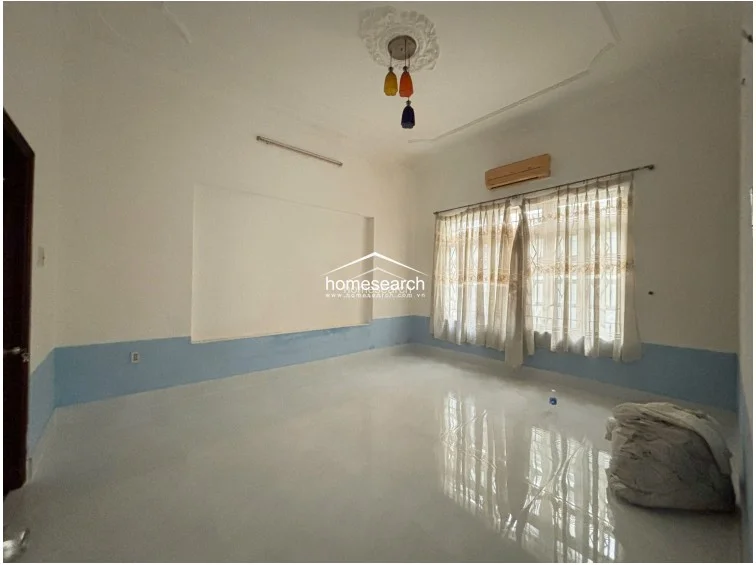 This spacious bedroom features high ceilings with decorative crown molding and a central ceiling medallion. The room is finished with light-colored tile flooring, white walls with a blue wainscot detail, and includes a wall-mounted air conditioning unit and a large window with sheer curtains for natural light.