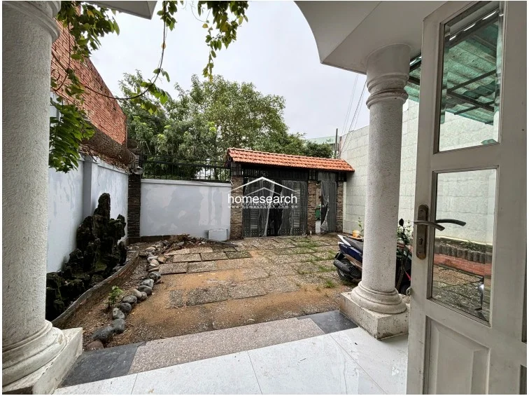 The front courtyard provides a private outdoor space featuring a stone-paved ground and a decorative rock feature against the side wall. The area is accessed via a gated entrance and connects directly to the main house through a white-framed glass door, offering a transition between the street and the interior.