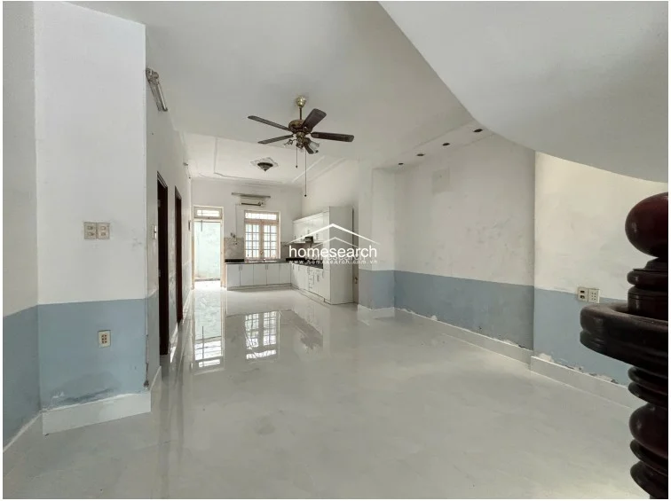 This open-plan space transitions from a living area into a kitchen equipped with white cabinetry and a wall-mounted AC unit. The room maintains a consistent aesthetic with white walls, blue wainscoting, and glossy tile flooring, and includes a wooden staircase railing in the foreground.