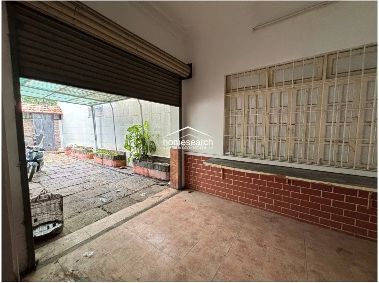 This semi-enclosed garage or multipurpose area features a roll-up metal door and a large window with security bars overlooking the courtyard. The space has simple tile flooring and a brick-accented wall, serving as a functional transition zone that connects the exterior driveway to the interior living quarters.