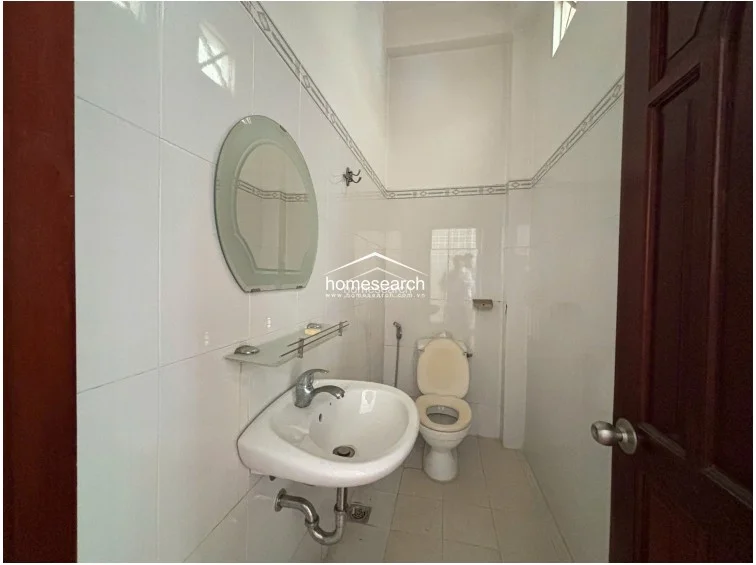 This compact bathroom is finished with white wall tiles and matching floor tiles, creating a clean and bright atmosphere. It is equipped with a pedestal sink, a wall-mounted mirror with a glass shelf, and a standard toilet, all maintained in a functional condition.