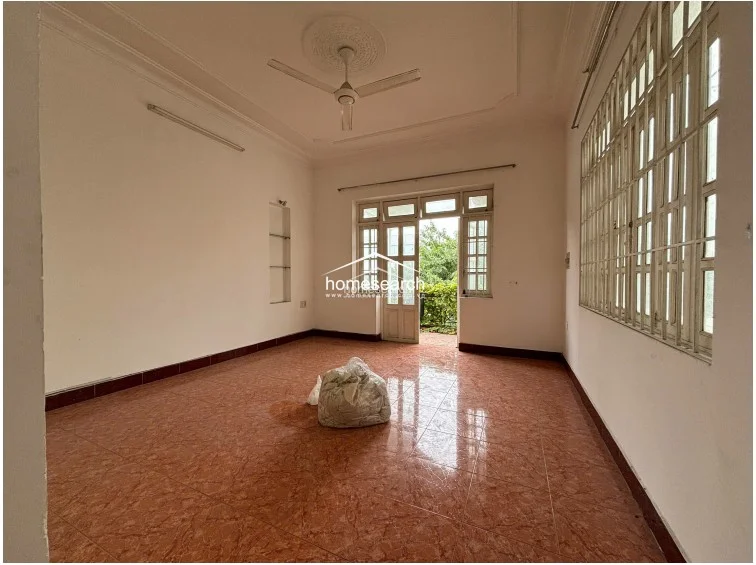 This spacious room is characterized by reddish-brown patterned tile flooring and white walls with a dark baseboard. It includes a ceiling fan, a large window with security bars, and a glass-paned door that leads to a balcony, offering a view of greenery.