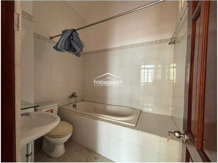 This bathroom is fully tiled in white, featuring a built-in bathtub with a handheld shower fixture. It includes a pedestal sink and a shower curtain rod, showing a functional, well-maintained layout with standard sanitary fixtures.