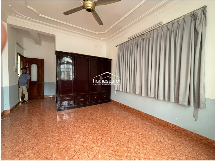 This view highlights a large, dark wood wardrobe providing substantial storage, set against a backdrop of light blue lower wall accents and white upper walls. The room is equipped with a ceiling fan and a large window covered by grey curtains, maintaining the consistent tile flooring seen throughout the property.