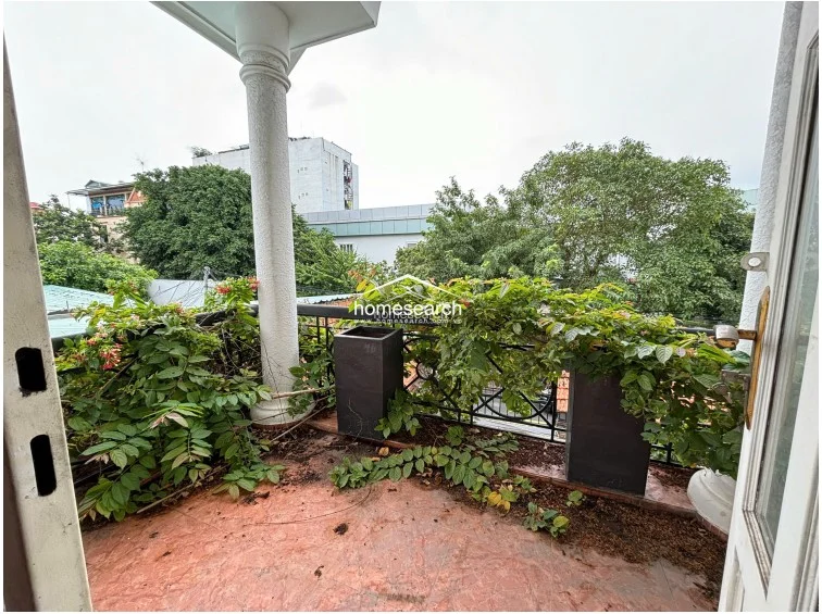 The private balcony features a classic architectural column and metal railings, overlooking a lush, green neighborhood view. While the space offers a pleasant outdoor retreat, the balcony floor and planters currently show signs of neglect with significant debris and overgrown vegetation that require cleaning and maintenance.