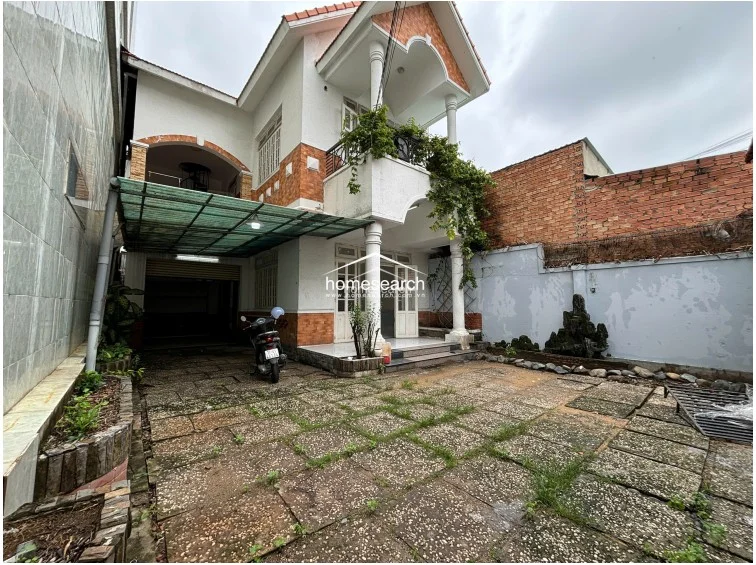 This two-story detached home features a prominent front courtyard with stone tiling and a covered carport area. The exterior showcases a white stucco finish with decorative orange brick accents, a second-floor balcony with climbing greenery, and classic columns, though the overall condition shows signs of weathering and requires maintenance.