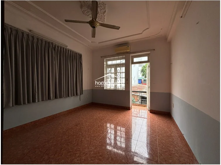 This angle emphasizes the room's layout, showing the connection between the interior living space and the outdoor balcony area. The space is functional, featuring a ceiling fan, air conditioning, and a neutral color palette, though it appears to require some maintenance regarding the window security grilles and general upkeep.