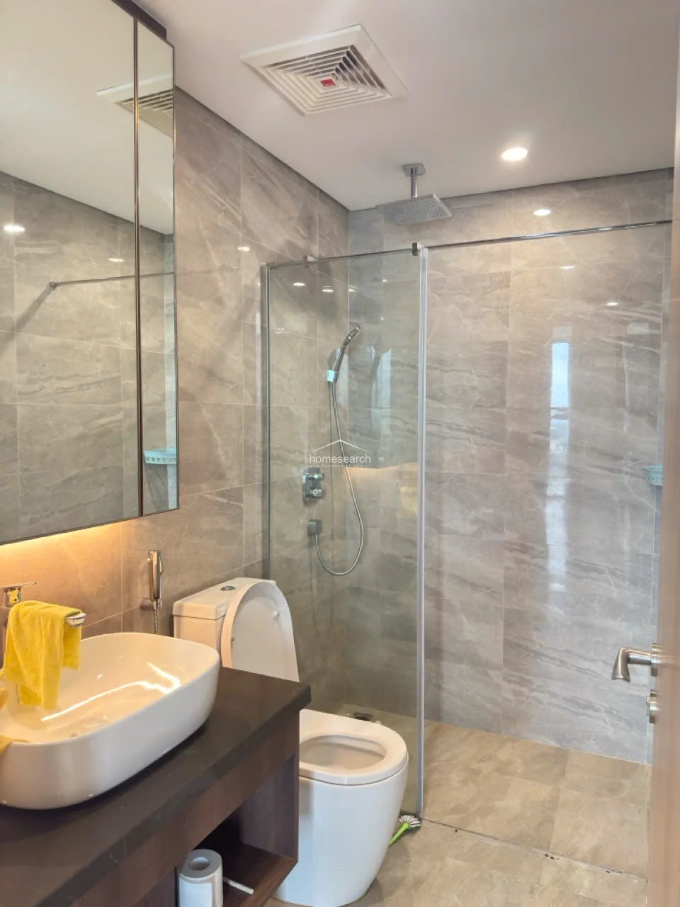 This compact, well-maintained bathroom utilizes a glass-enclosed walk-in shower with a rainfall showerhead and a separate handheld sprayer. The vanity features a vessel sink on a dark stone countertop, complemented by a large mirrored cabinet and recessed ceiling lighting. The neutral, earth-toned tiling throughout creates a cohesive and polished look.