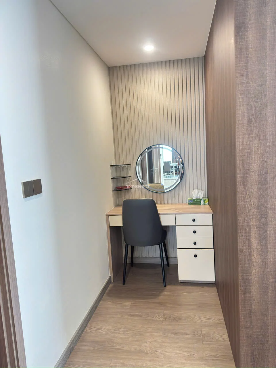 This dedicated vanity area is tucked into a functional alcove, featuring a sleek desk with integrated drawers and a stylish circular mirror. The space is accented with vertical slat paneling and floating glass shelves, offering a refined and organized spot for personal grooming.