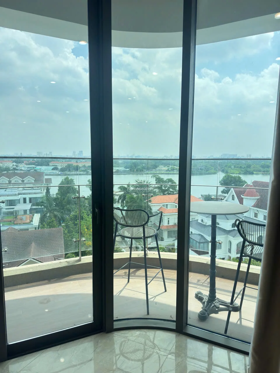 This image shows a curved, private balcony featuring a small bistro set, perfect for enjoying the expansive river views. The space is enclosed by a modern glass railing, ensuring safety without compromising the scenery, and is finished with neutral-toned tiling. The floor-to-ceiling glass doors provide a seamless transition from the interior to this outdoor relaxation area.