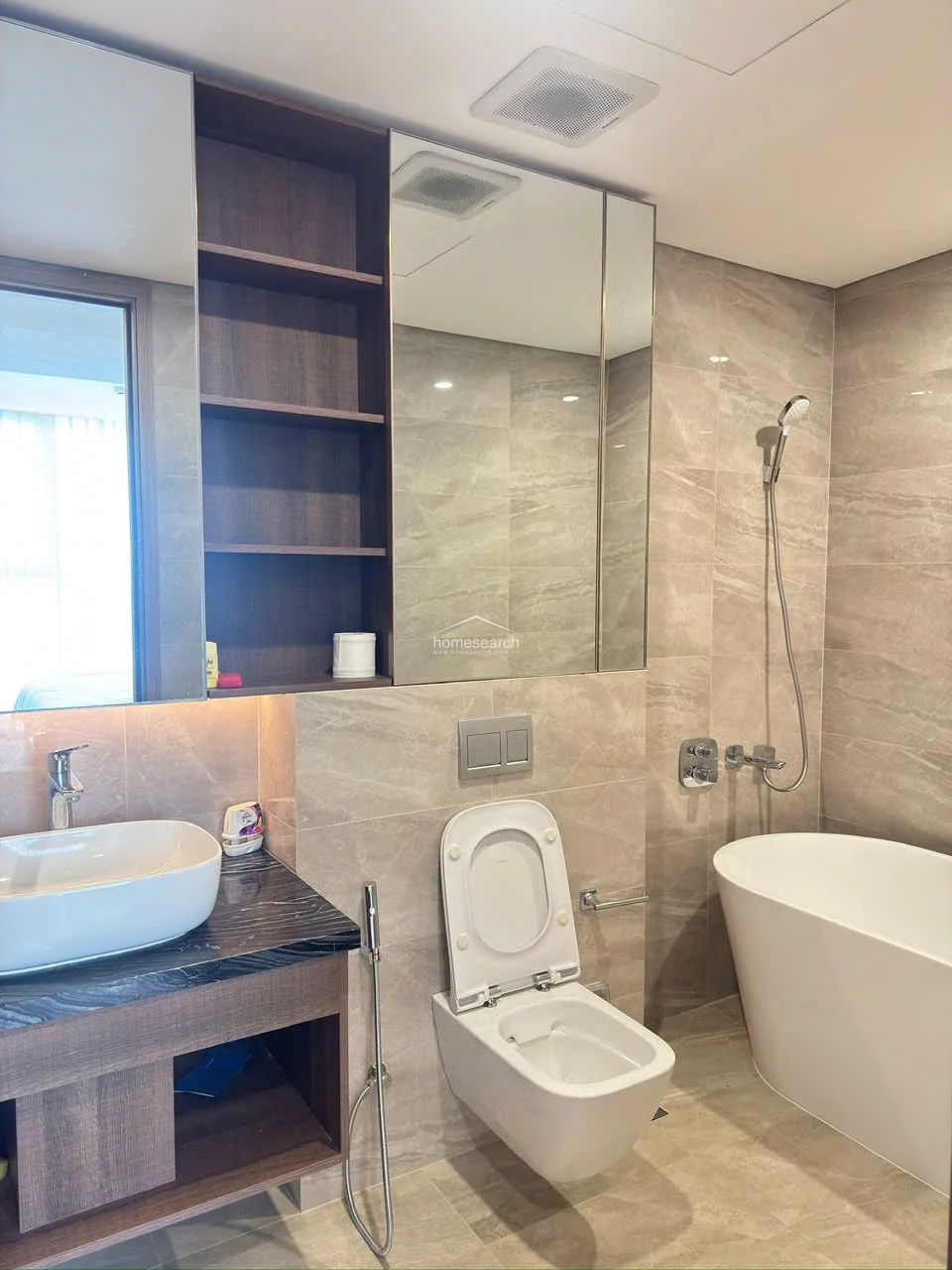 This modern bathroom features a sleek, freestanding soaking tub paired with a wall-mounted toilet and a vessel sink set on a dark stone vanity. The space is finished in neutral, large-format beige tiles and includes integrated open shelving and a mirrored medicine cabinet for storage. The overall condition is excellent, reflecting a contemporary and high-end design aesthetic.