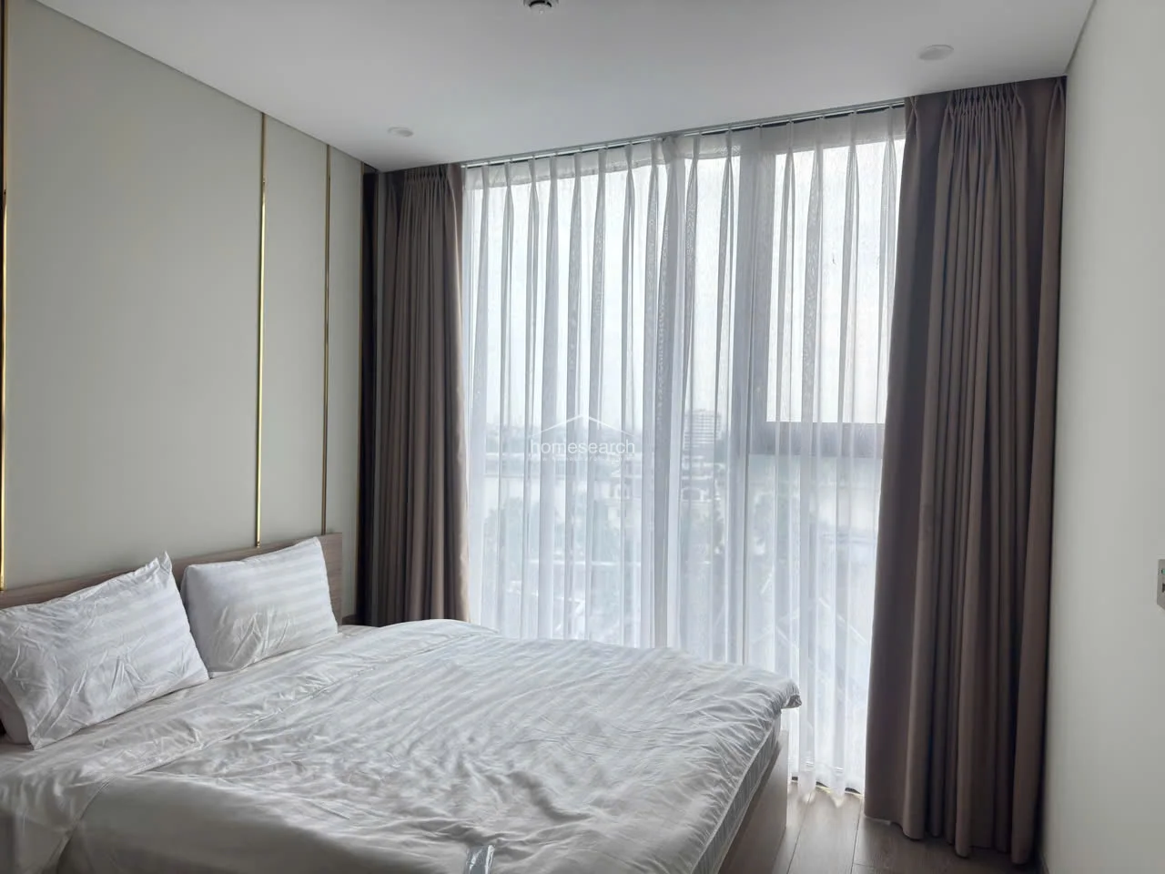 This bedroom features a modern, minimalist design with a queen-sized bed and a large floor-to-ceiling window dressed in layered sheer and heavy taupe curtains. The room is finished with light wood-look flooring and a feature wall accented with vertical gold trim, creating a sophisticated and clean aesthetic.