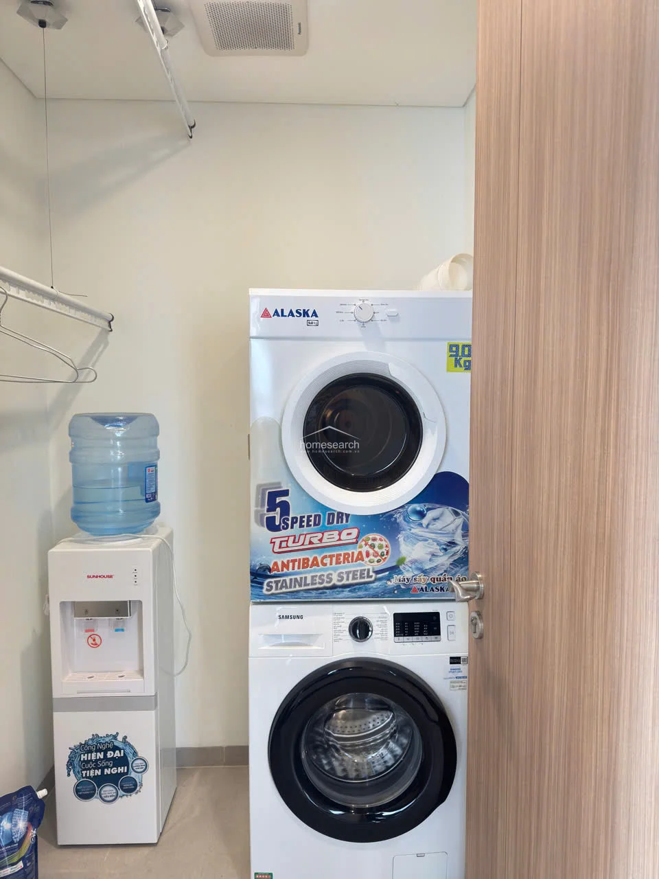 This dedicated laundry/utility space is efficiently organized with a stacked washer and dryer unit, maximizing the small footprint. It includes a water dispenser and a wall-mounted rack for hanging clothes, finished with clean, light-colored walls and wood-toned doors. The area is well-maintained and provides a practical, hidden solution for household chores.