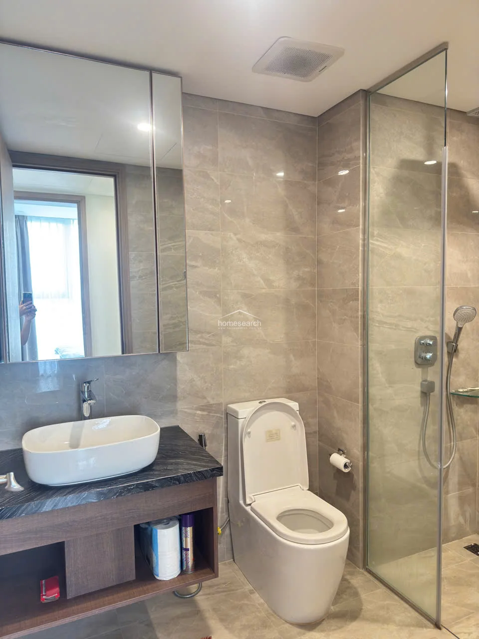 This bathroom layout is nearly identical to the previous one, featuring a glass-partitioned shower area and a modern vessel sink vanity. The room is finished with consistent beige wall and floor tiles, providing a clean, uniform appearance. It includes essential fixtures like a wall-mounted toilet, a mirrored medicine cabinet, and efficient ceiling ventilation.