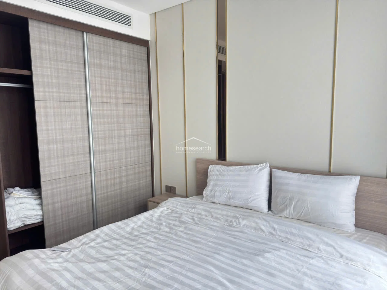 This view highlights the bedroom's functional storage, featuring a built-in floor-to-ceiling sliding wardrobe with a textured, light wood finish. The room is equipped with a concealed ceiling-mounted AC unit, ensuring a sleek look while maintaining a comfortable climate.