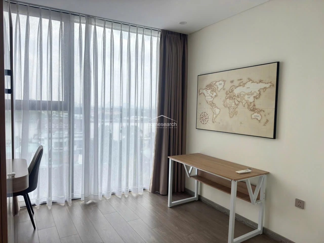 This room serves as a bright, minimalist home office or study area, featuring wood-look flooring and soft, neutral wall tones. It is bathed in natural light from a large window dressed with elegant, layered curtains and includes a functional desk setup and a world map wall decoration. The space feels calm and productive, maintaining a clean and modern aesthetic throughout.