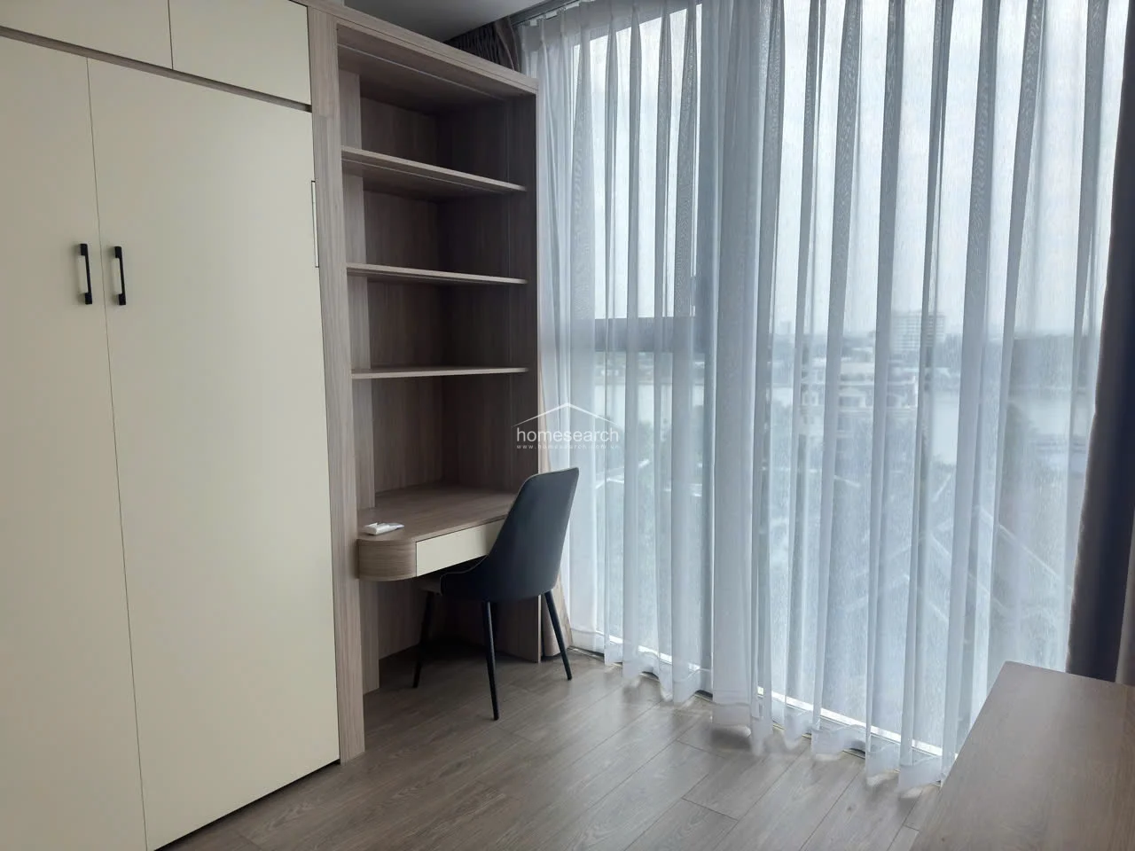 This functional bedroom or study area is equipped with a built-in wardrobe and an integrated desk with open shelving for storage. The room features wood-look flooring and floor-to-ceiling windows covered by sheer curtains, which allow for plenty of natural light while maintaining privacy. The furniture is cohesive and modern, reflecting a high level of maintenance and thoughtful space planning.