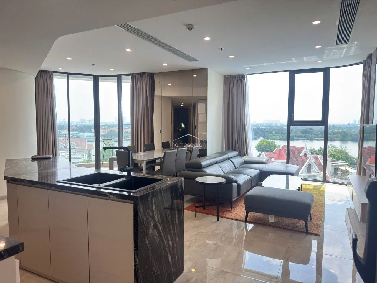 This open-concept living and dining area features a sleek, modern kitchen with a dark marble-topped island and high-gloss white cabinetry. The space is bathed in natural light from floor-to-ceiling curved windows that offer panoramic river views, complemented by polished light-toned tile flooring and recessed ceiling lighting.