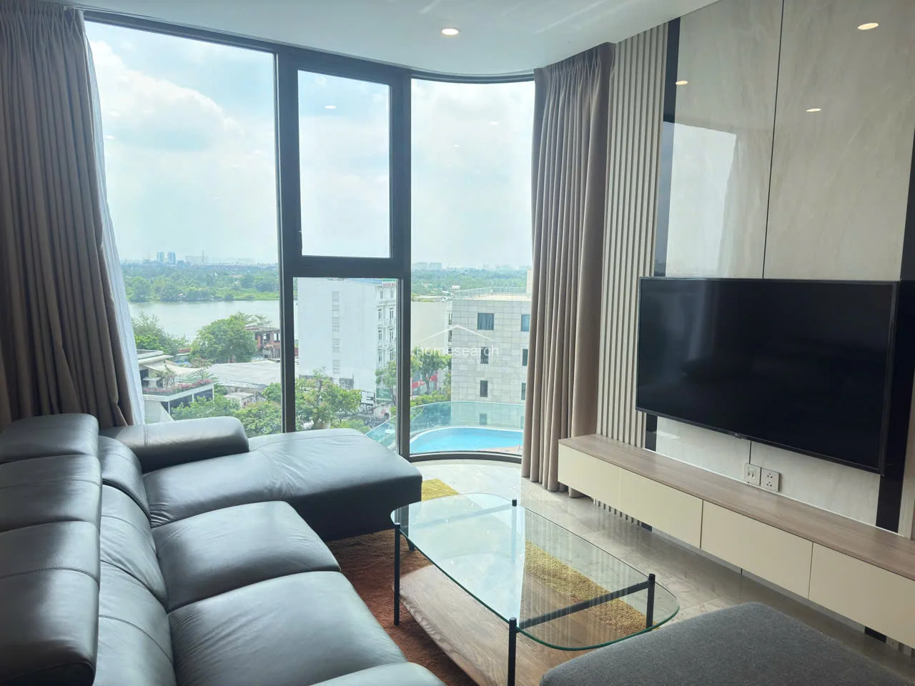 The media wall features a custom floating console and wall-mounted television, accented by vertical wood slats and marble-effect paneling. The curved window design frames a stunning view of the river and a glimpse of a nearby swimming pool, emphasizing the property's high-end, modern architectural style.