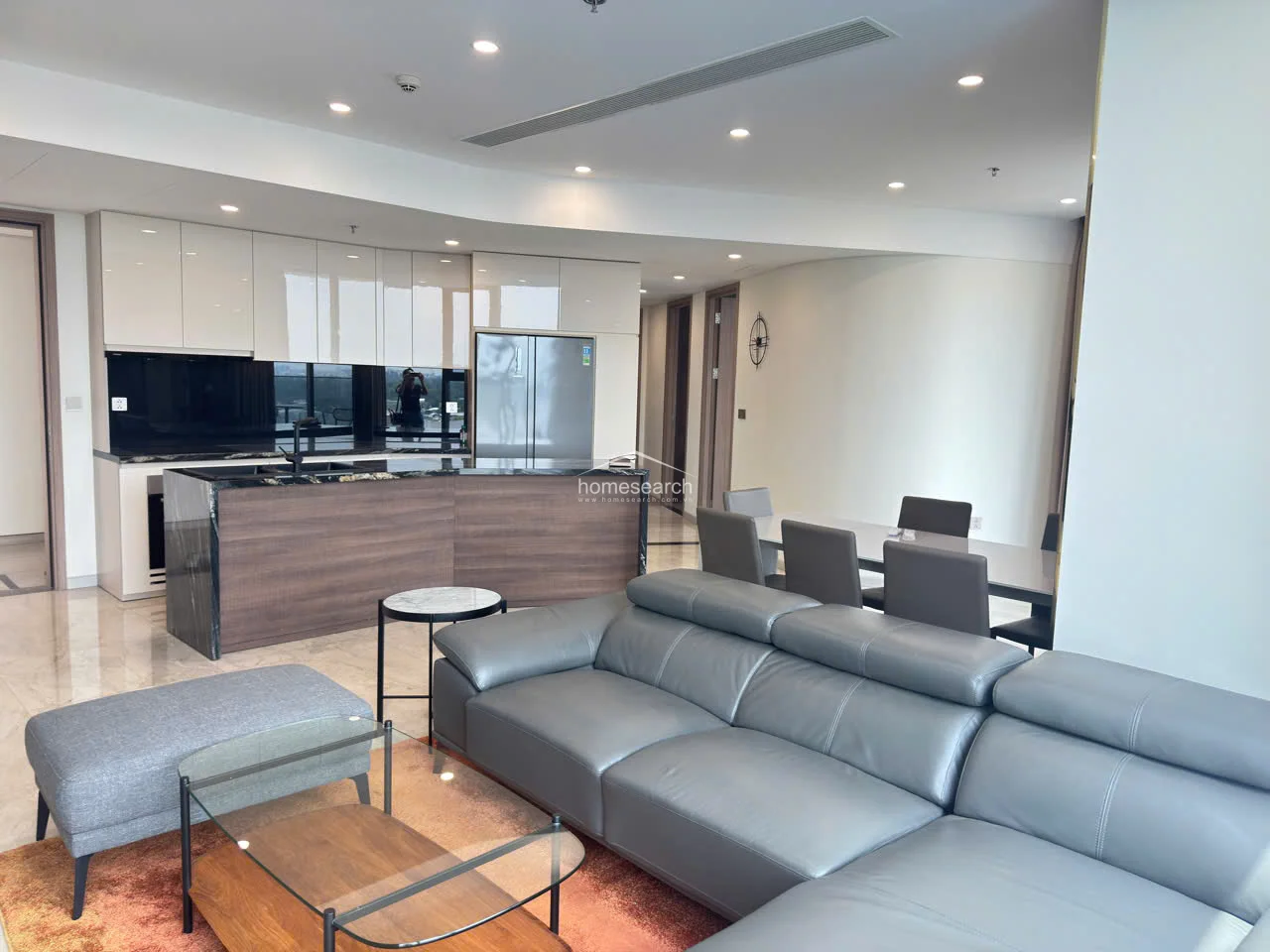 This view highlights the kitchen's functional layout, showcasing a large stainless steel refrigerator and ample storage within the minimalist white upper cabinets. The dark wood-paneled island provides a sophisticated contrast to the light tile floors, while the integrated ceiling air conditioning ensures a clean, uncluttered aesthetic throughout the living space.
