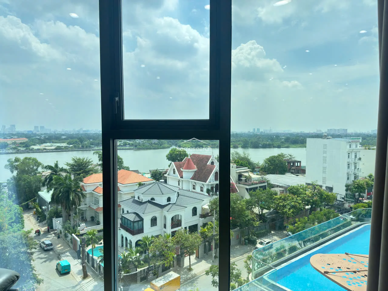 This image captures a stunning, unobstructed view of a river and surrounding residential neighborhood through a large, modern floor-to-ceiling window. The perspective highlights the building's height, looking down onto a private swimming pool and lush greenery below. The window frame is sleek and dark, emphasizing the clean, contemporary architectural style of the property.