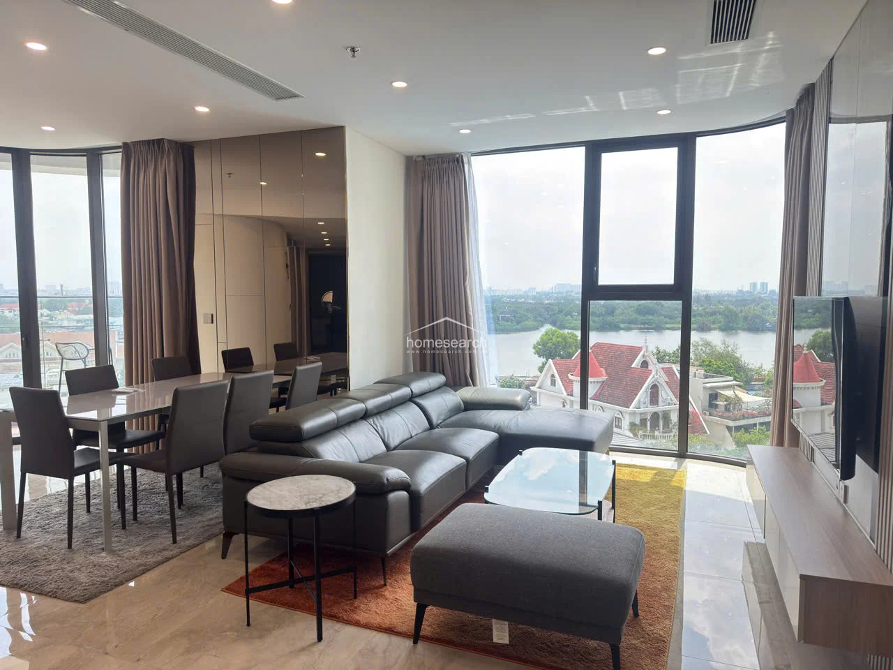 The living room is anchored by a large, contemporary grey leather sectional and a glass-top coffee table, set against a backdrop of neutral walls and floor-to-ceiling drapery. The layout flows seamlessly into the dining area, which is mirrored by a wall of tinted glass panels that enhance the sense of depth and luxury in the room.