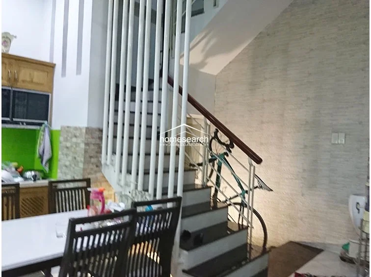 This image shows a kitchen and dining area featuring a prominent staircase with white railings and dark treads. The kitchen is equipped with wooden cabinetry and a vibrant green backsplash, while the flooring consists of light-colored tiles. The space appears well-maintained with an open-plan layout that flows directly into the dining area.