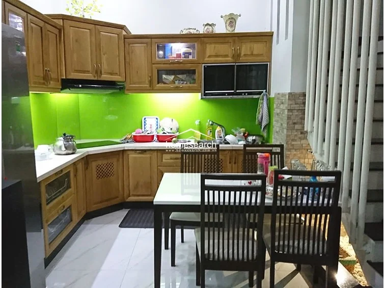 This view highlights the kitchen layout, which utilizes an L-shaped wooden cabinetry design and a bright green glass backsplash. The dining area is seamlessly integrated, featuring a white table and matching wooden chairs positioned near the staircase. The overall condition is clean and functional, with ample storage provided by the overhead and base cabinets.