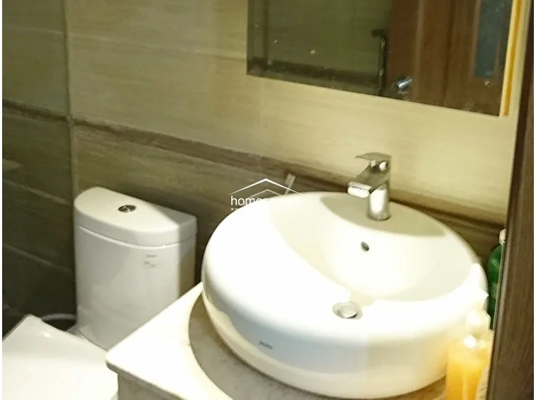 The bathroom is equipped with a modern, white vessel sink set upon a stone vanity and a standard flush toilet. The walls are finished with neutral-toned, horizontal-patterned tiles, creating a clean and functional aesthetic.