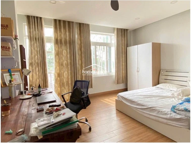 This bedroom features warm wood-toned flooring and a large window dressed with floor-to-ceiling beige curtains, allowing for ample natural light. The room is furnished with a comfortable bed, a white wardrobe, and a large wooden desk, providing a balanced space for rest and productivity.