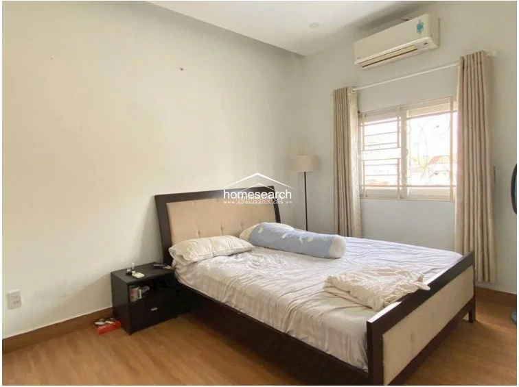 This bedroom is finished with light-colored walls and wood-patterned flooring, giving it a bright and airy feel. It is equipped with a wall-mounted AC unit, a dark wood bed frame with a padded headboard, and a window with security bars and beige curtains.