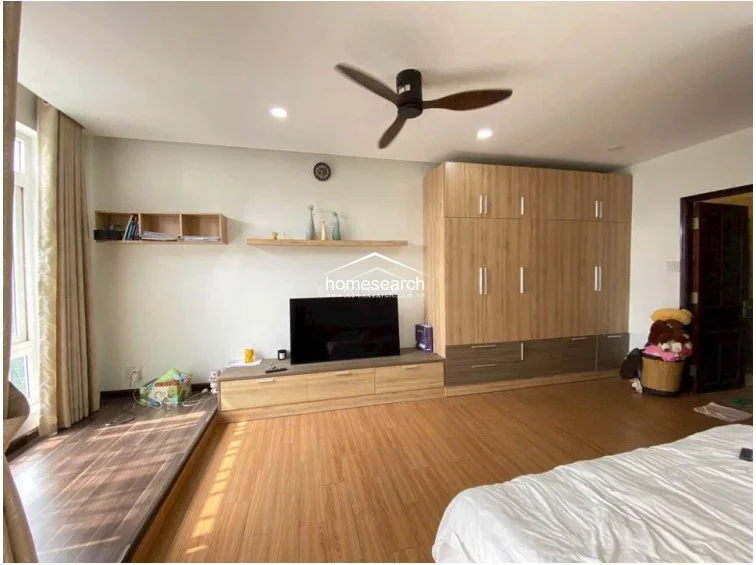 This bedroom features a warm, wood-toned aesthetic with matching laminate flooring and a large, built-in floor-to-ceiling wardrobe. The room is well-lit by a large window area that includes a raised platform, providing a functional space for seating or storage.