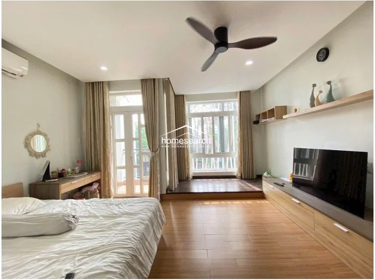 The bedroom layout emphasizes natural light through a glass door leading to a balcony and a large window, both dressed in neutral curtains. It is equipped with a wall-mounted AC unit, a modern ceiling fan, and a dedicated vanity area, maintaining a clean and well-maintained appearance.